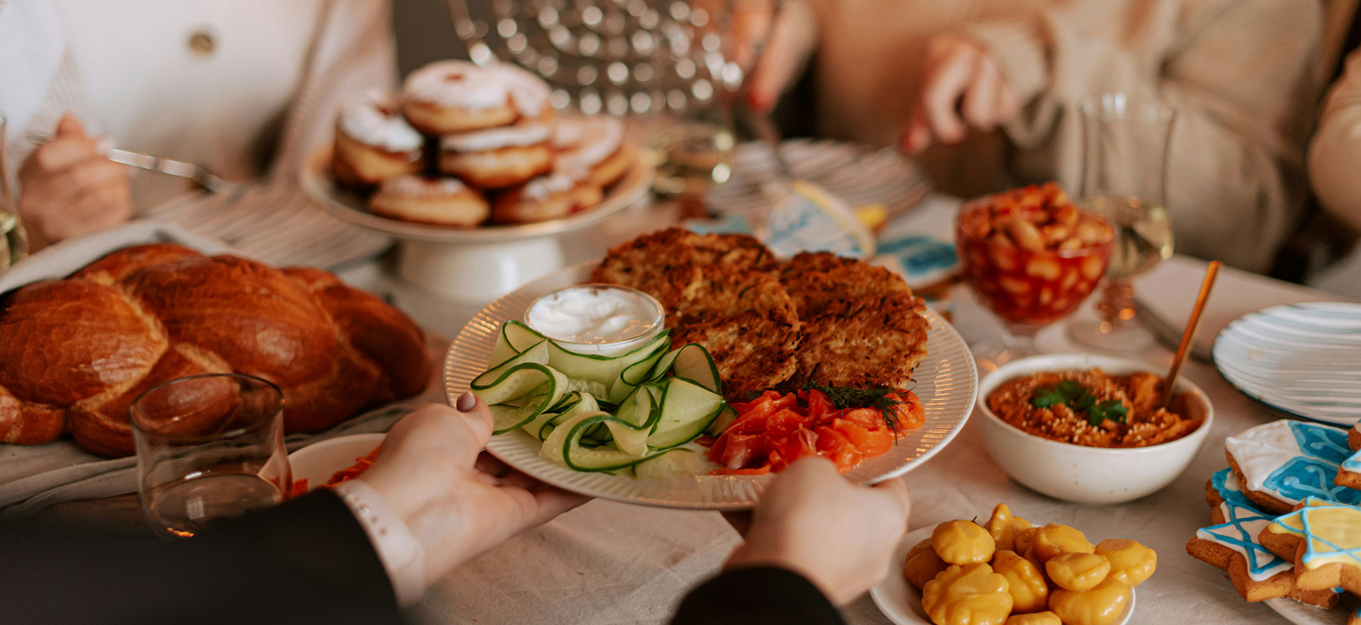 Ein gedeckter Tisch mit verschiedenen Speisen. Eine Hand reicht einen Teller mit gebratenen Kartoffelpuffern, Gurken, Tomaten und einer Schale mit Sahne. Im Hintergrund sind Gebäck und andere Snacks sichtbar. Die Atmosphäre wirkt festlich und einladend.