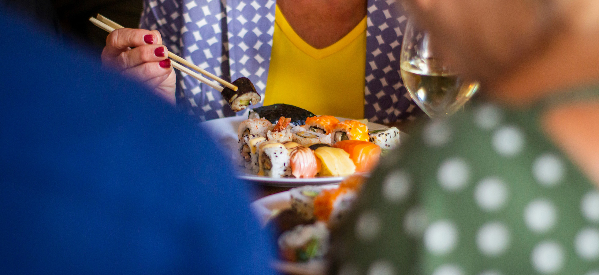 Eine Hand mit roten Nägeln hält Stäbchen und greift nach einem Sushi-Stück auf einem Teller. Im Hintergrund sind verschwommene Gestalten und ein Glas Wein zu sehen. Der Fokus liegt auf dem Sushi und der Hand, die es aufnimmt.