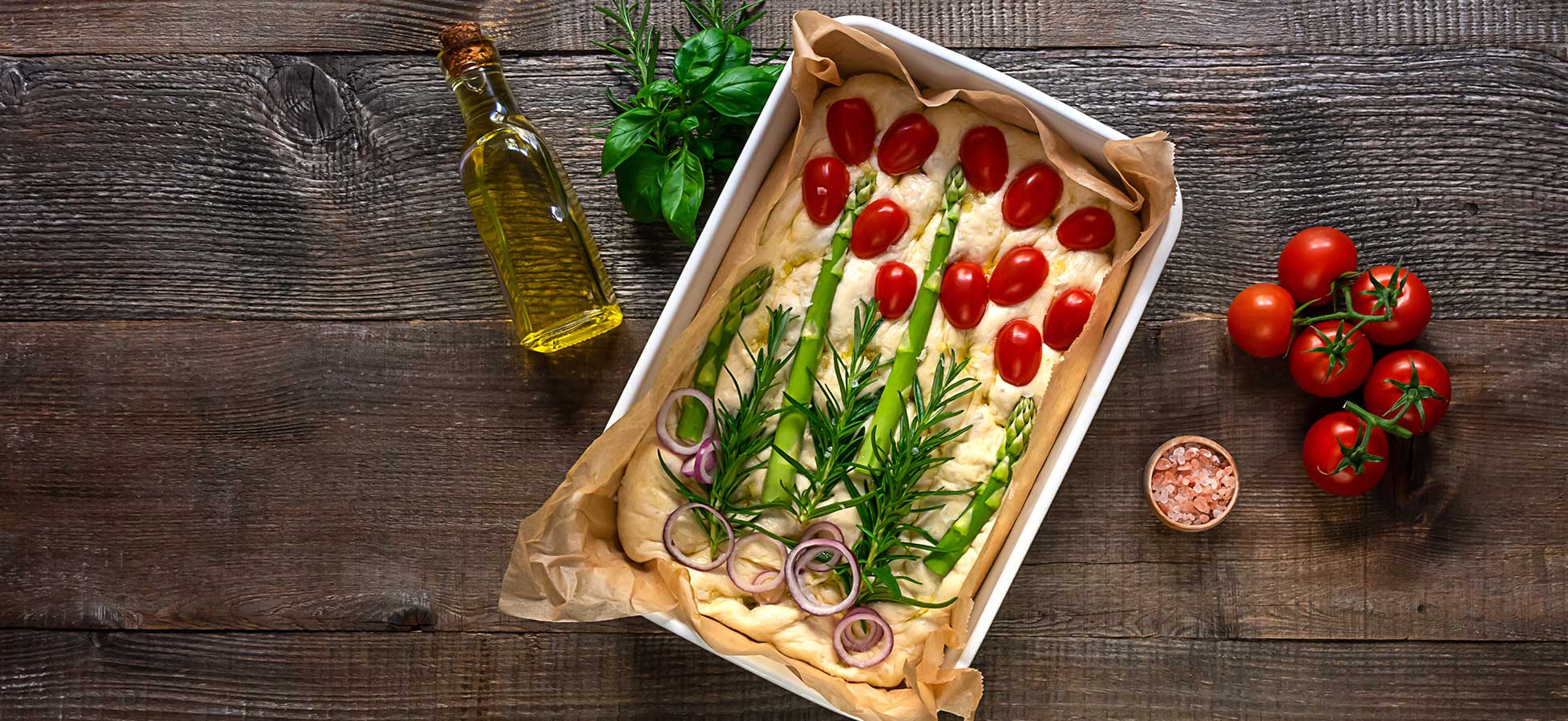 Ein Backblech mit rohem Focaccia-Teig, belegt mit Kirschtomaten, Rosmarin, Frühlingszwiebeln und roten Zwiebelscheiben auf einem Holztisch. 