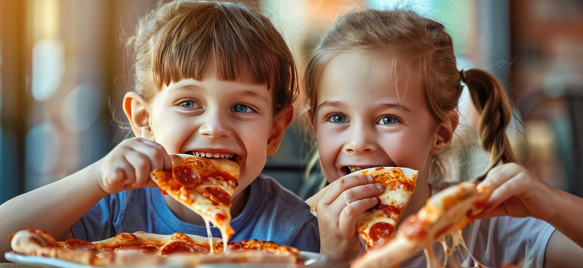 Zwei Kinder sitzen lächelnd an einem Tisch und essen Stücke einer Peperonipizza.