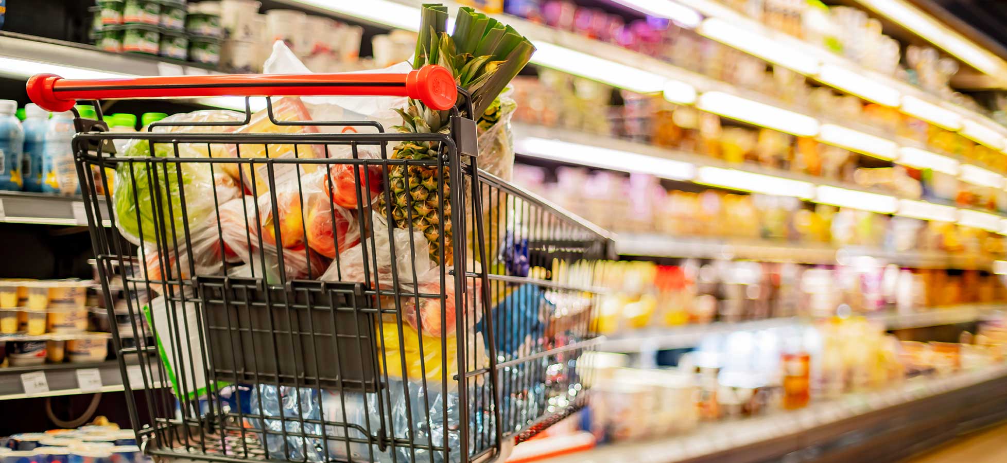 Ein Einkaufswagen voller Lebensmittel, darunter eine Ananas, in einem Supermarktgang mit Regalen voller verschiedener Produkte.