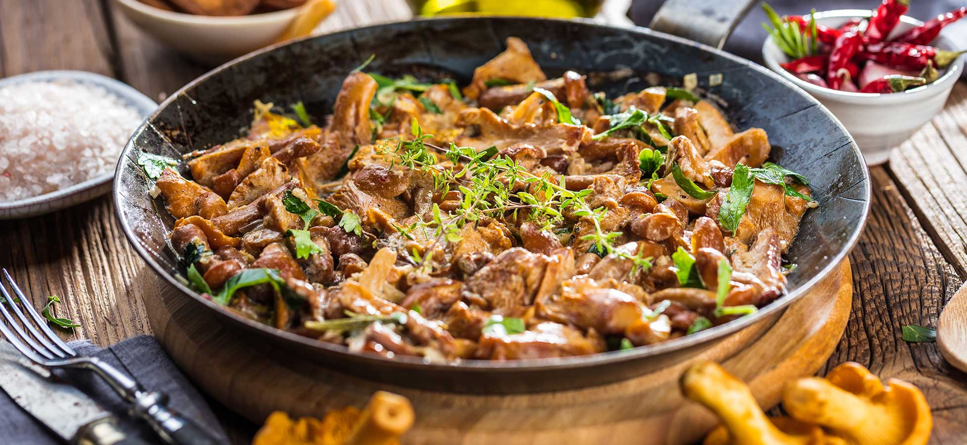 Eine Bratpfanne gefüllt mit einem cremigen Pilzgericht, garniert mit frischen Kräutern, auf einem Holztisch mit verschiedenen Zutaten platziert.