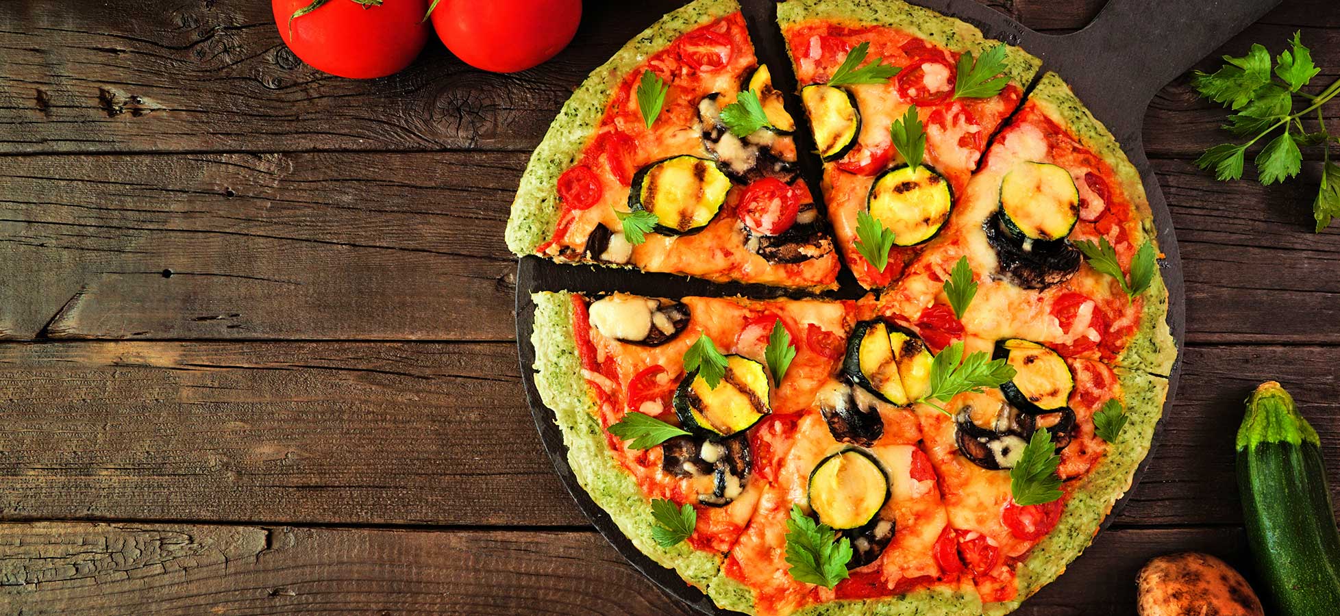 Eine Gemüsepizza mit Zucchini, Tomaten, Champignons und Petersilie auf einem Holztisch, teilweise angeschnitten. 