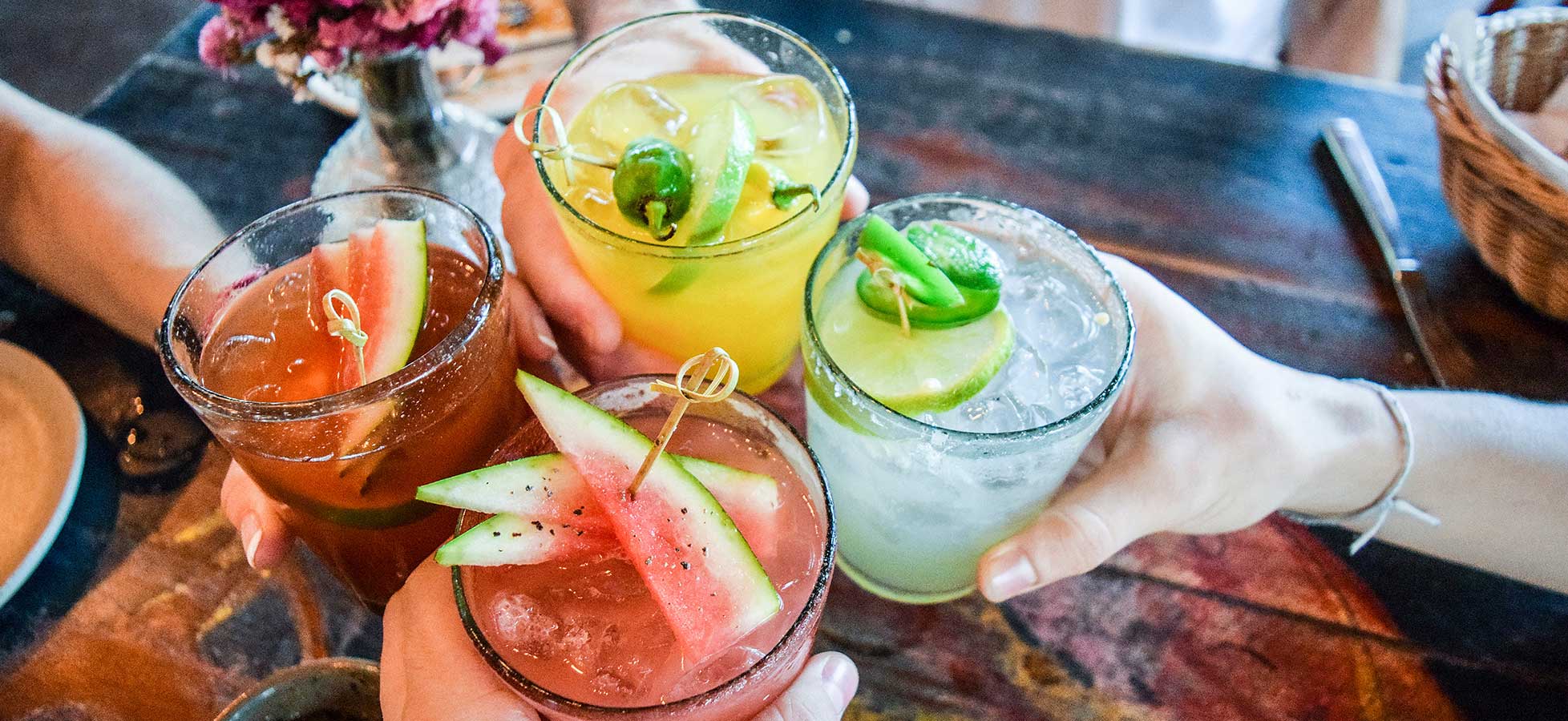 Vier Hände heben Gläser mit bunten, garnierten Cocktails vor einem dunklen Holztisch mit einem Korb und einem Blumengesteck.