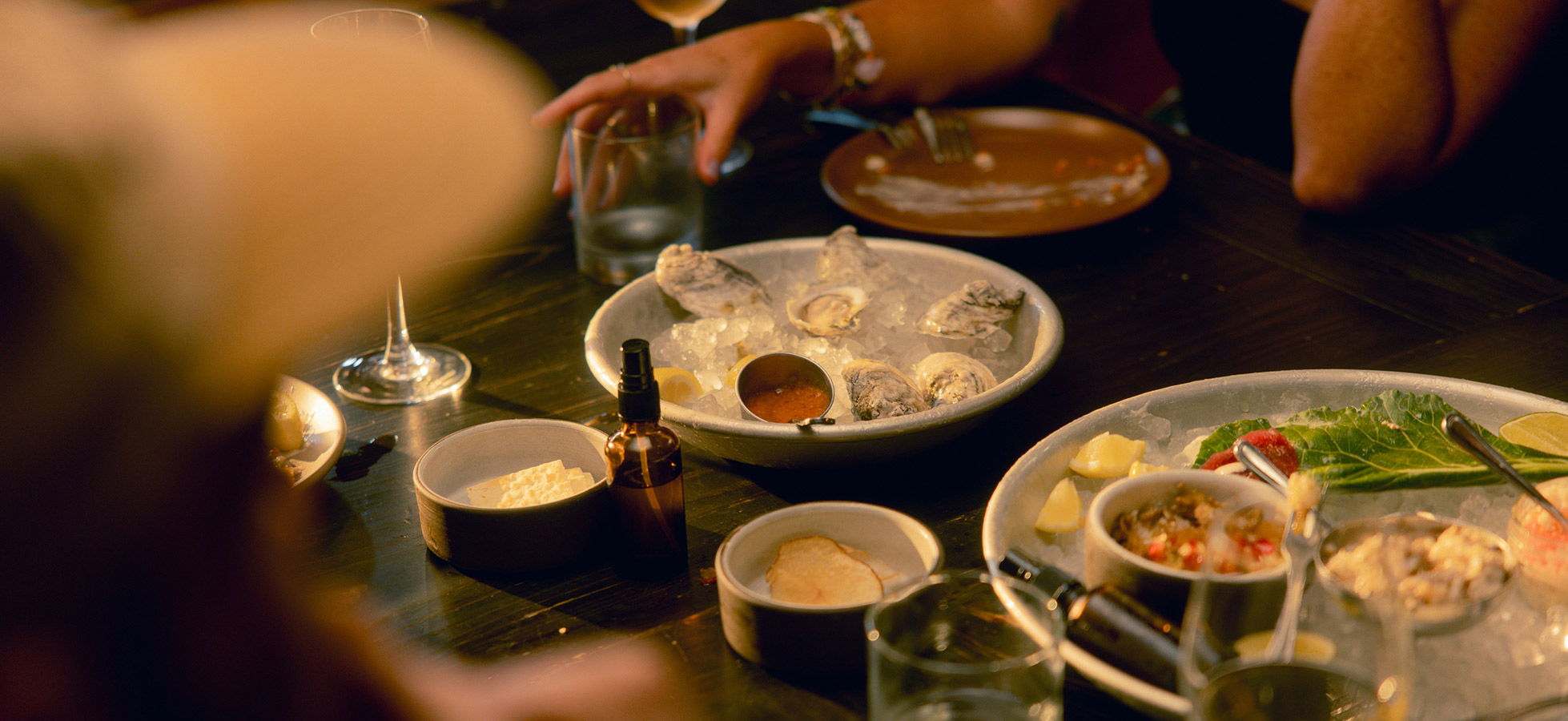 Ein Tisch mit verschiedenen Gerichten, darunter frische Austern auf Eis, sowie kleine Schalen mit Soßen und Beilagen. Im Hintergrund sind Gläser und eine Hand, die nach einer Speise greift. Das Ambiente wirkt gesellig und einladend.