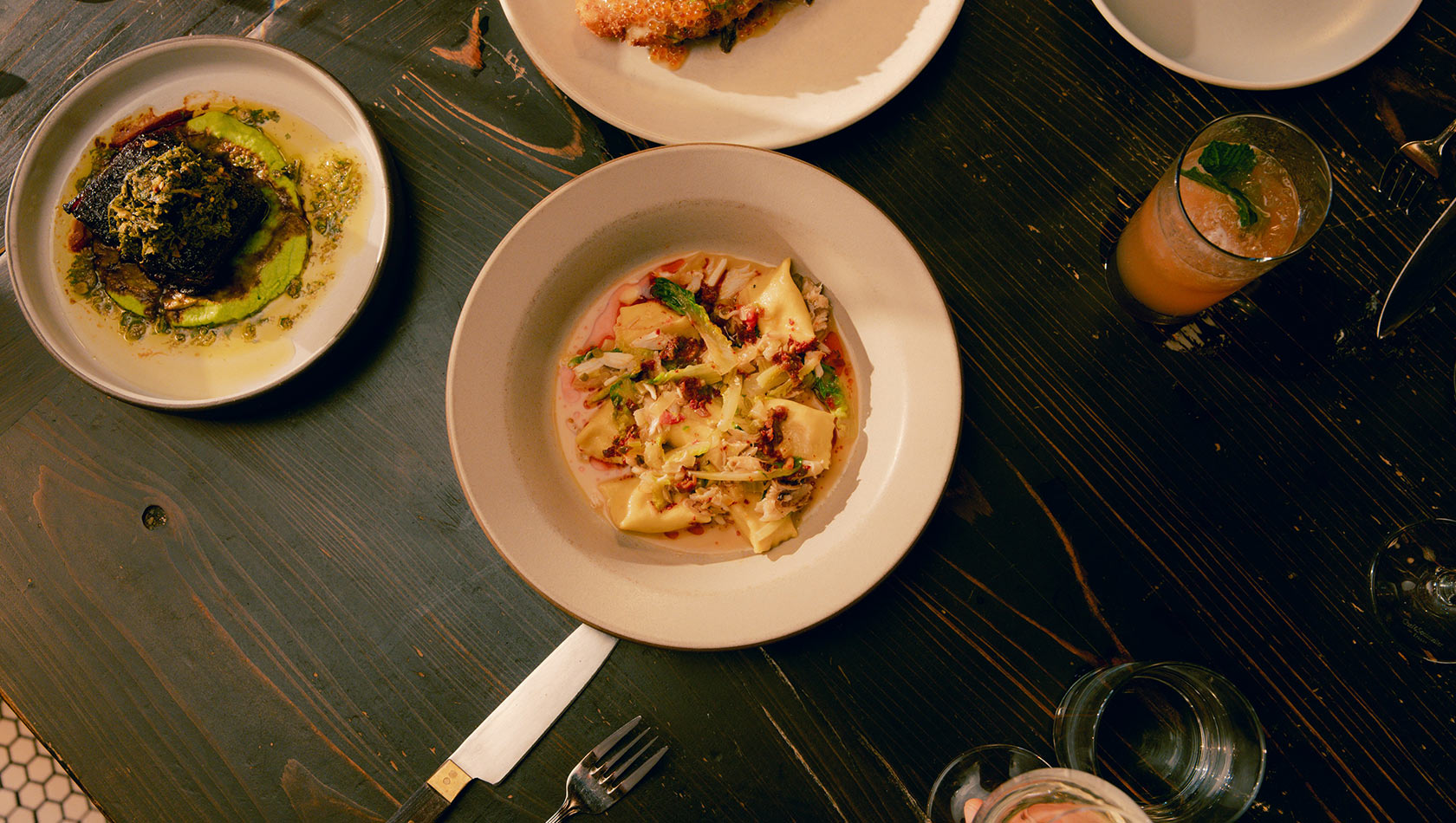 Ein Teller mit Ravioli in einer cremigen Sauce, garniert mit frischem Gemüse und rotem Dressing, befindet sich in der Mitte eines Holztisches. Nebenan liegt ein weiterer Teller mit einem dunklen Gericht und einer grünen Sauce. Auch ein Getränk mit Minze ist sichtbar.