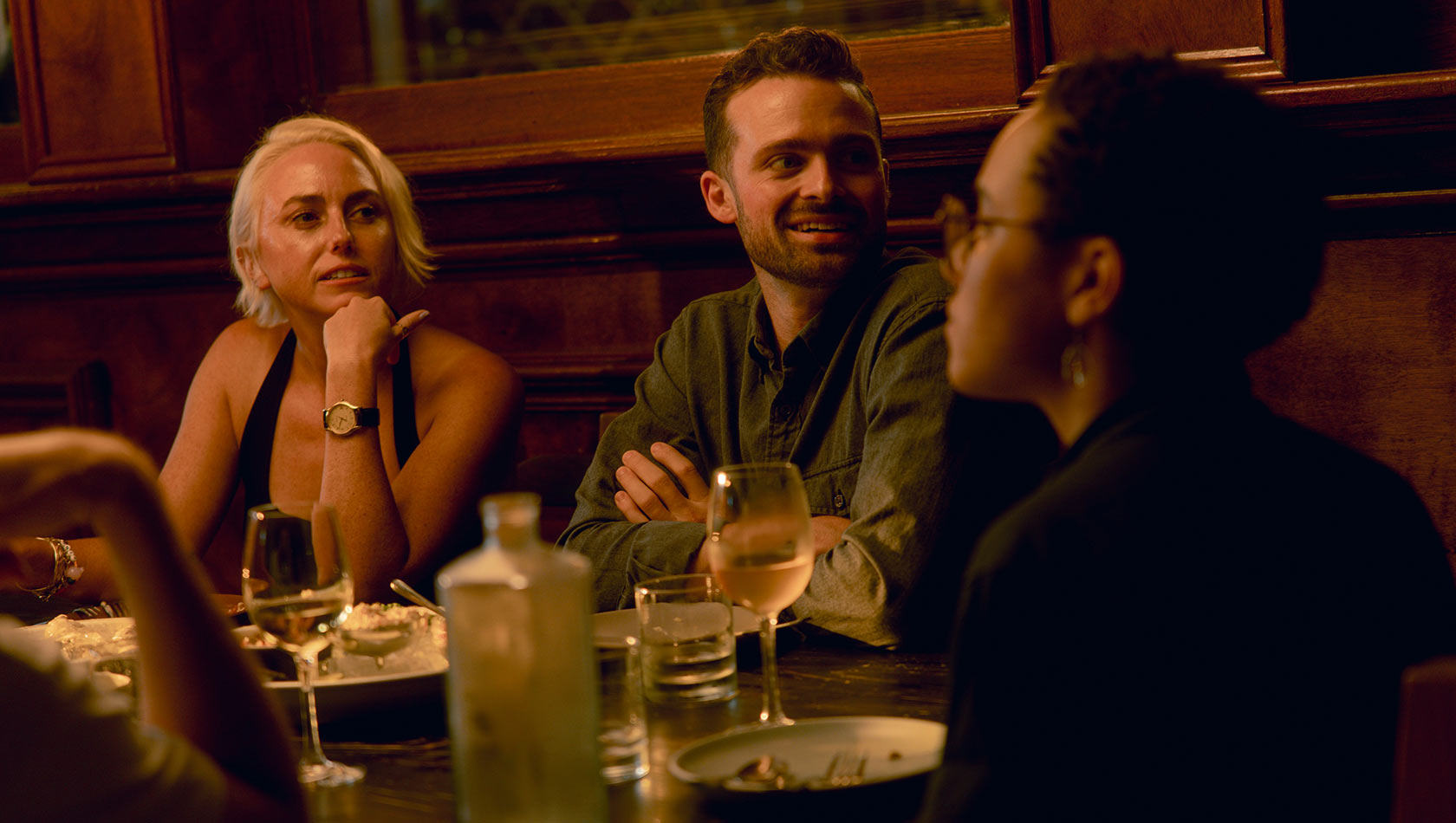 Eine Gruppe von drei Personen sitzt an einem Tisch in einem Restaurant. Eine Frau mit blonden Haaren schaut nachdenklich, während ein Mann in einem grauen Hemd lächelt und mit ihr spricht. Eine weitere Frau mit kurzen, lockigen Haaren hört aufmerksam zu. Getränke und Essen sind auf dem Tisch.