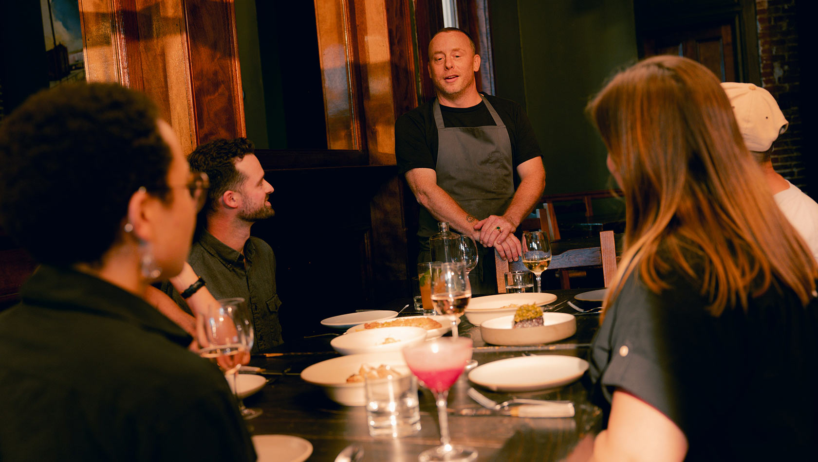 Ein Koch steht an einem Tisch, umgeben von Gästen, die aufmerksam zuhören. Auf dem Tisch sind mehrere Schalen mit Essen und Getränke platziert. Das Ambiente ist gemütlich, und die Gäste scheinen am Gespräch interessiert zu sein.