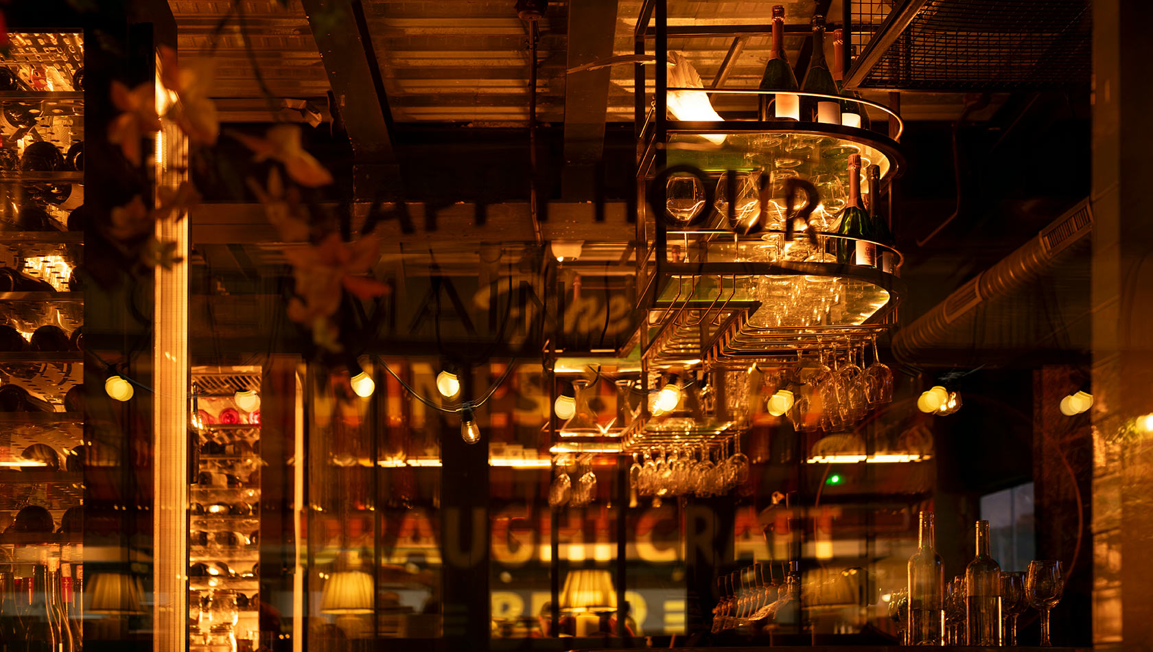 Das Bild zeigt eine stilvolle Bar mit warmem Licht. Im Vordergrund hängt ein elegantes Lichtobjekt, das den Schriftzug "Happy Hour" trägt. Um das Licht sind Flaschen und Gläser angeordnet, während hinter dem Tresen eine Auswahl an Getränken sichtbar ist.