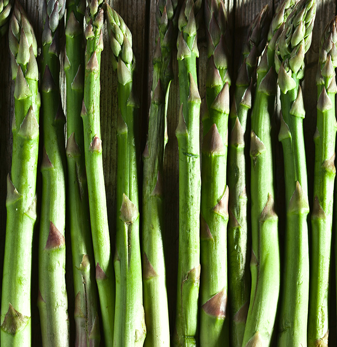 Eine Reihe von frischen grünen Spargelstangen, die auf einem Holztisch liegen. Die Stangen sind gleichmäßig geschnitten und weisen typische knospige Spitzen auf. Die Farbe ist leuchtend grün, was auf Frische hinweist.