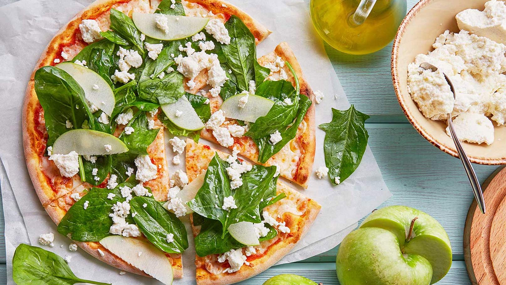 Das Bild zeigt eine kreative Variante von Pizza, die appetitlich angerichtet ist. Neben der farbenfrohen Pizza, die mit verschiedenen Zutaten belegt ist, steht ein Glas Olivenöl, das einen Hauch von mediterranem Flair verleiht. Daneben befindet sich eine Schüssel mit cremigem Crème fraîche, perfekt zum Dippen oder als zusätzliche Füllung. Frische Äpfel runden das Arrangement ab und bieten einen fruchtigen Kontrast zur herzhaften Pizza. 