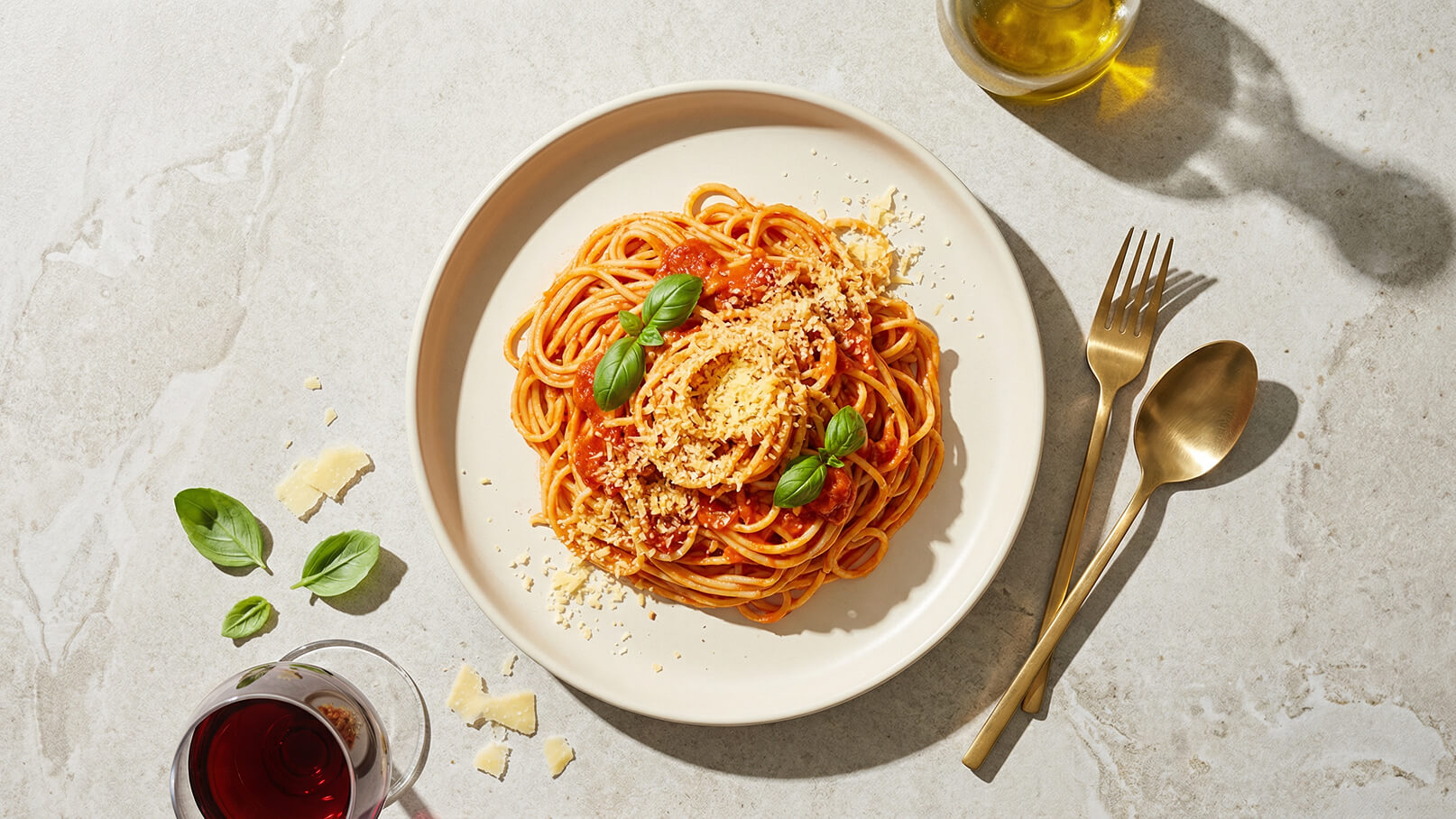 Ein Teller mit Spaghetti in Tomatensoße, garniert mit frischem Basilikum und geriebenem Käse. Daneben liegen eine Gabel und ein Löffel aus goldfarbenem Metall. Im Hintergrund ist ein Glas Rotwein sichtbar.