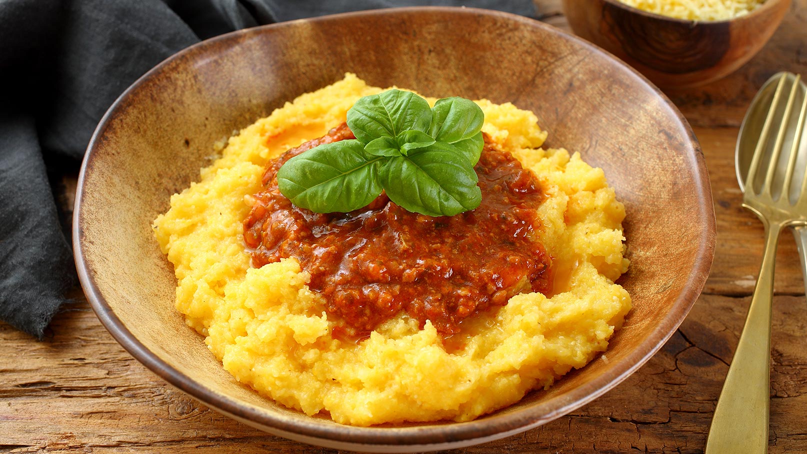 Auf dem Bild ist eine braune Schüssel zu sehen, gefüllt mit dampfender Polenta, die großzügig mit einer köstlichen Paradeisersoße bedeckt ist. Das Gericht ist mit frischen Basilikumblättern dekoriert, die dem Ganzen eine frische und aromatische Note verleihen. Die braune Schüssel unterstreicht die rustikale und hausgemachte Atmosphäre des Gerichts. 