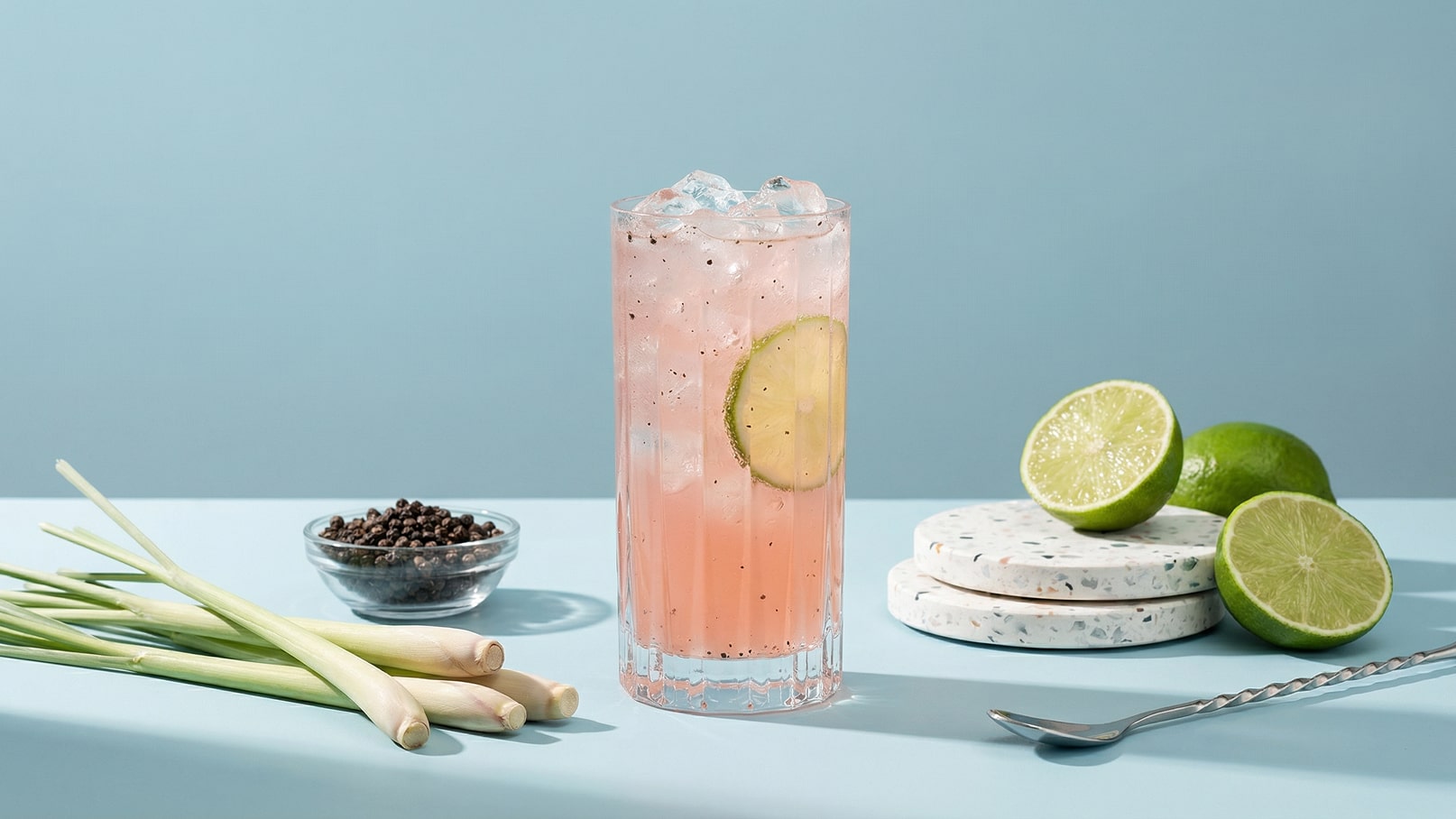 Ein erfrischender pinker Cocktail mit Eiswürfeln und einer Limettenscheibe ist im Vordergrund zu sehen. Daneben liegen frische Zitronengrasstängel, eine Schale mit Pfefferkörnern und halbierte Limetten auf Untersetzern. Der Hintergrund ist in einem sanften Blau gehalten.