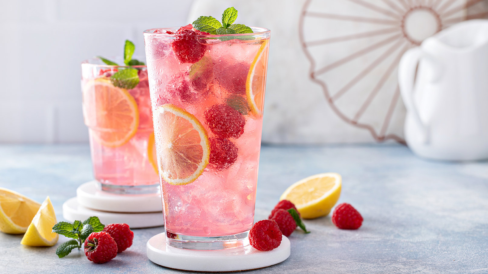 Auf dem Bild sind zwei Gläser zu sehen, randvoll gefüllt mit einem leckeren Himbeercocktail, der mit frischen Himbeeren, Zitronen, Minze und Eiswürfeln verziert ist. Die Gläser stehen auf weißen Steinuntersetzern, die das Bild elegant wirken lassen. Neben den Gläsern liegen frische Himbeeren, Minze und Zitronen, die die frischen Zutaten des Cocktails betonen. 