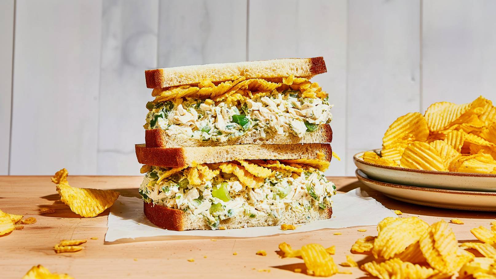 Ein hoher Sandwich-Turm besteht aus zwei Scheiben Brot, gefüllt mit einer Mischung aus gehacktem Hähnchen, Kräutern und einer Schicht Kartoffelchips. Neben dem Sandwich liegt ein Teller mit weiteren Kartoffelchips. Der Hintergrund ist schlicht gehalten.