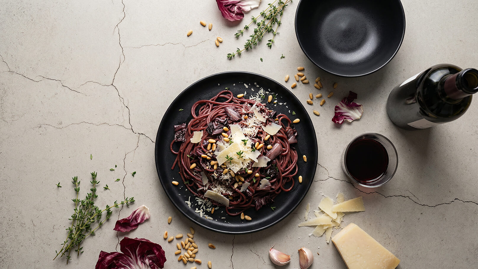 Ein schwarzer Teller mit tiefroten Spaghetti, garniert mit Rote-Bete-Blättern, geriebenem Käse und Pinienkernen. Um den Teller herum liegen frische Kräuter, eine Flasche Rotwein und ein Glas Rotwein. Der Hintergrund ist aus hellem, rissigem Stein.