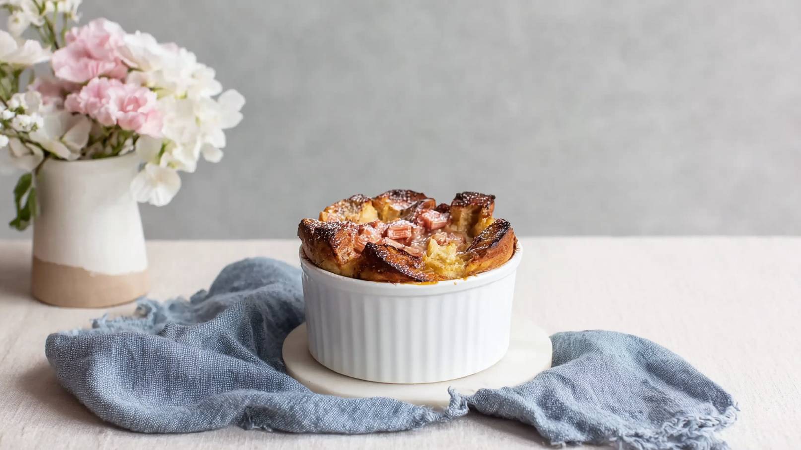 Eine helle, runde Auflaufform mit einer goldbraunen, knusprigen Oberseite steht auf einem weißen Untersetzer. Umgeben ist sie von einem hellen Tuch. Im Hintergrund ist eine Vase mit zarten, bunten Blumen zu sehen.