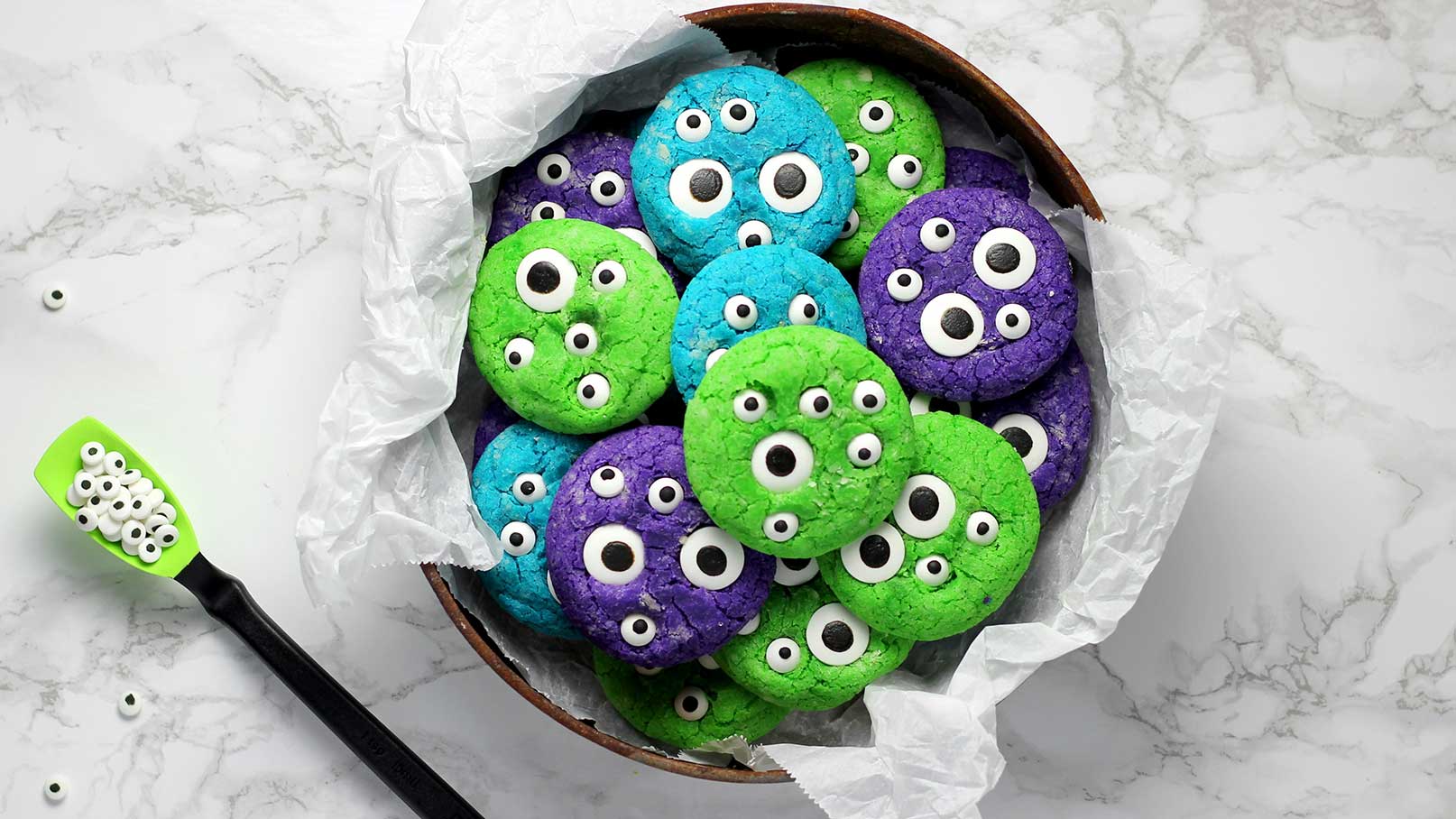 Eine Schüssel mit bunten, monsterähnlichen Keksen in den Farben Grün, Blau und Lila, die mit essbaren Augenkonturen verziert sind. Die Kekse sind übereinander gestapelt und wirken verspielt und einladend. Ein grüner Backspatel mit zusätzlichen Augenkonturen ist daneben platziert.