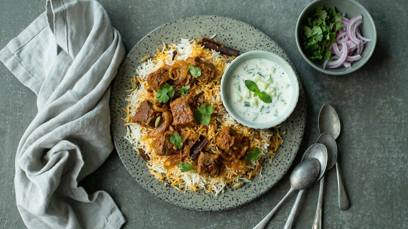 Ein Teller mit duftendem Biryani bestehend aus gewürztem Reis, zartem Fleisch und Zwiebeln, garniert mit frischem Koriander. Neben dem Hauptgericht befindet sich eine Schale mit Joghurt-Dip, der mit Gurkenstücken und einem Minzblatt dekoriert ist.