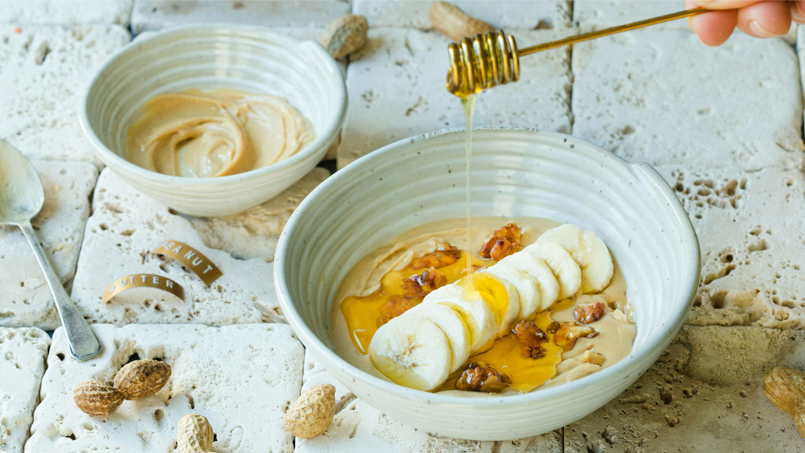 Eine Schüssel mit einer cremigen Erdnussbutter-Mischung, belegt mit Bananenscheiben und Nüssen. Honig wird darüber gegossen. Im Hintergrund steht eine zweite Schüssel mit Erdnussbutter. Die Szenerie ist auf einer hellen, steinernen Oberfläche arrangiert.