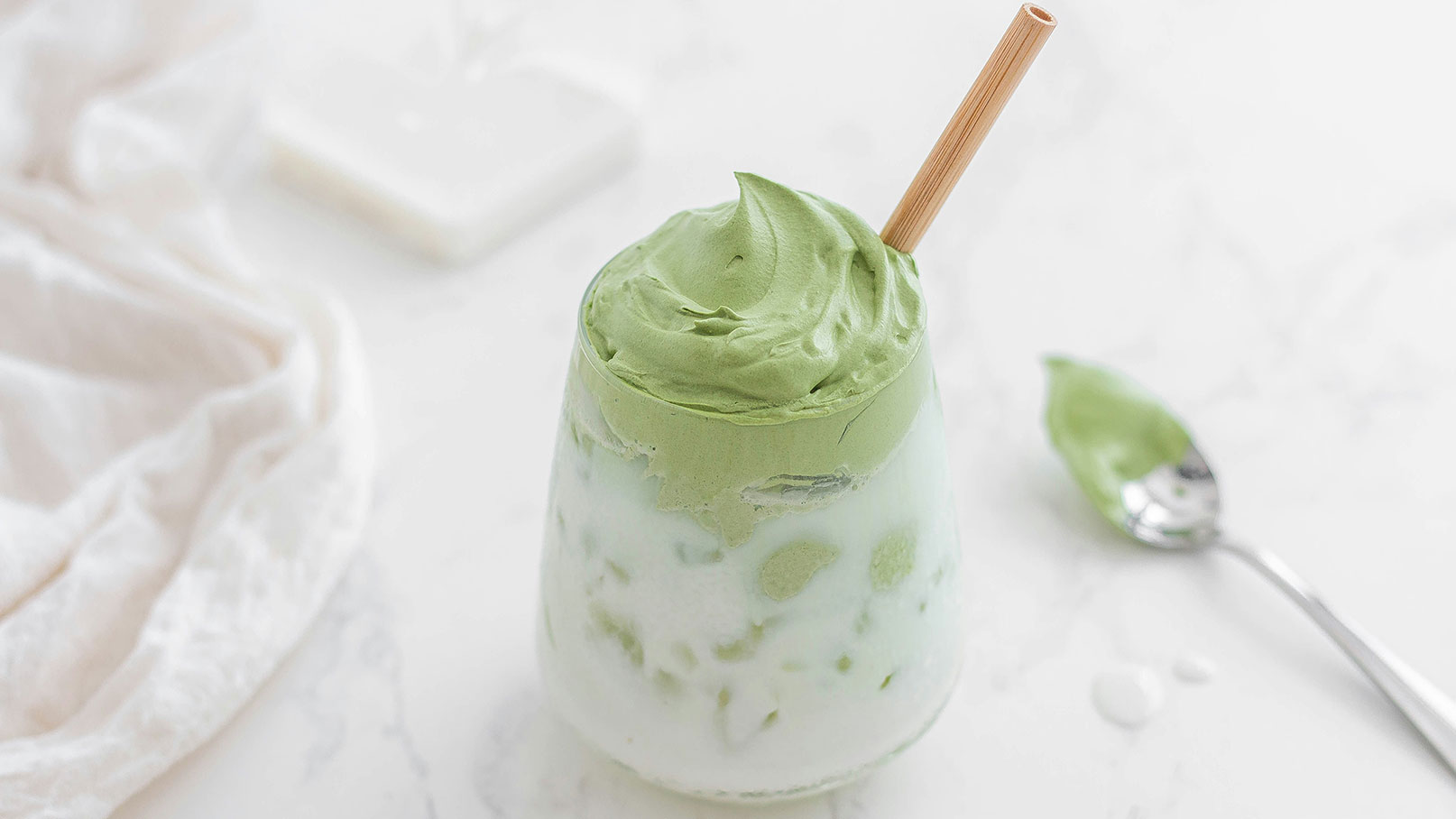 Ein Glas mit einer geschichteten grünen Creme, oben mit einer lockeren, grünen Schicht. Ein Strohhalm aus Holz steckt in der Creme. Im Hintergrund liegt ein Löffel mit einer kleinen Menge der gleichen grünen Creme. Der Untergrund ist hell und schlicht.