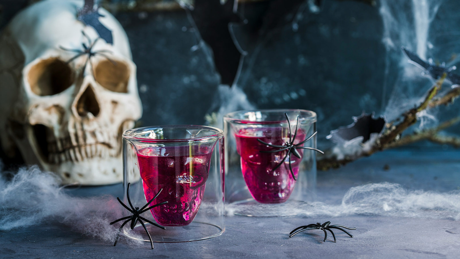 Zwei Gläser mit rotem Getränk, das in einer Totenschädelform gestaltet ist. Sie stehen vor einem Schädel und sind von schwarzen Spinnen und Spinnennetzen umgeben. Die Szene vermittelt eine gruselige Atmosphäre, passend zu Halloween.