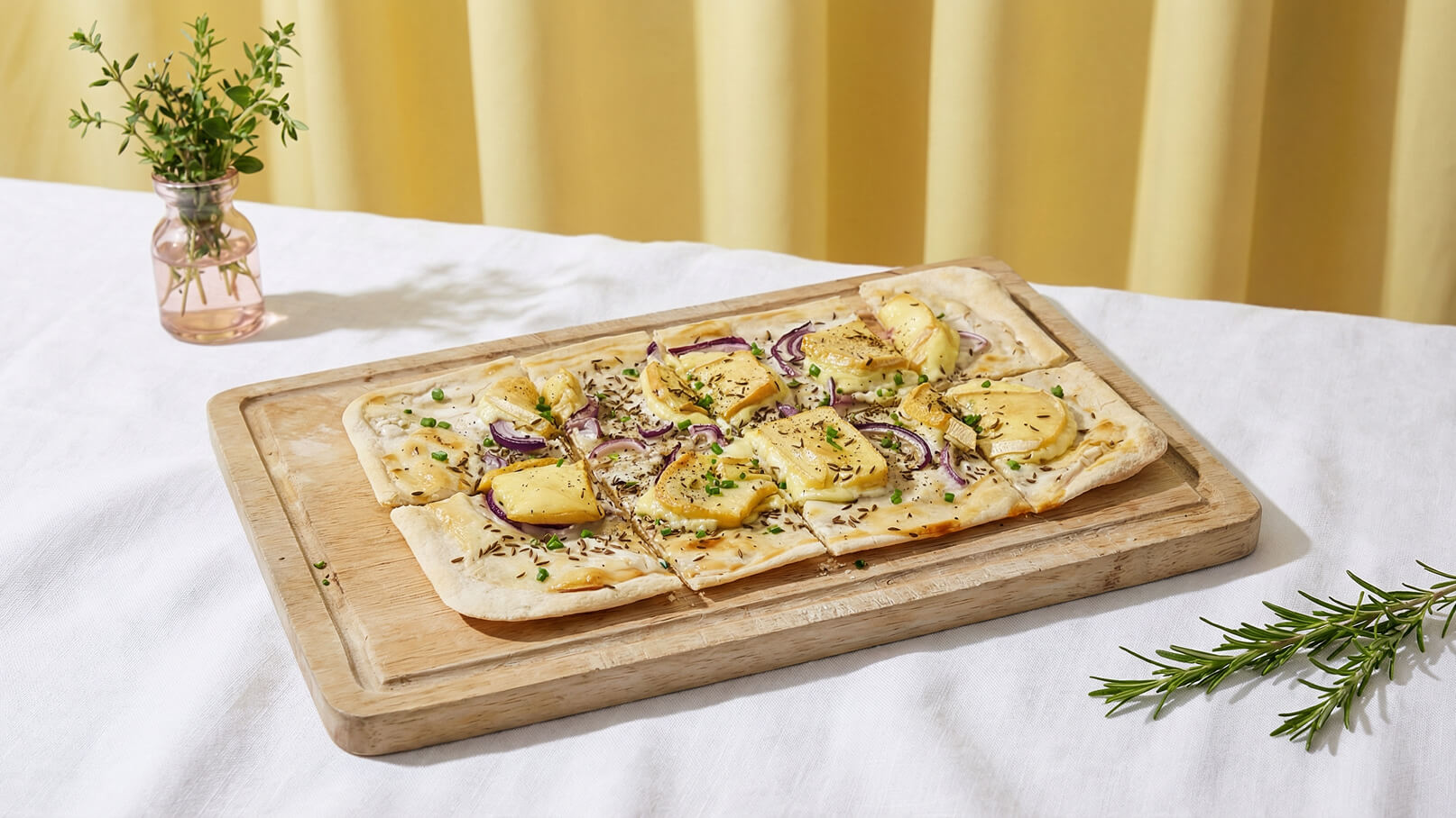 Eine Holzplatte mit einer rechteckigen Spezialität, belegt mit Zwiebeln und Kartoffelscheiben, steht auf einem weißen Tisch. Neben der Platte sind ein Kräutertopf und ein Rosmarinzweig platziert. Der Hintergrund ist gelblich und sorgt für eine warme Atmosphäre.