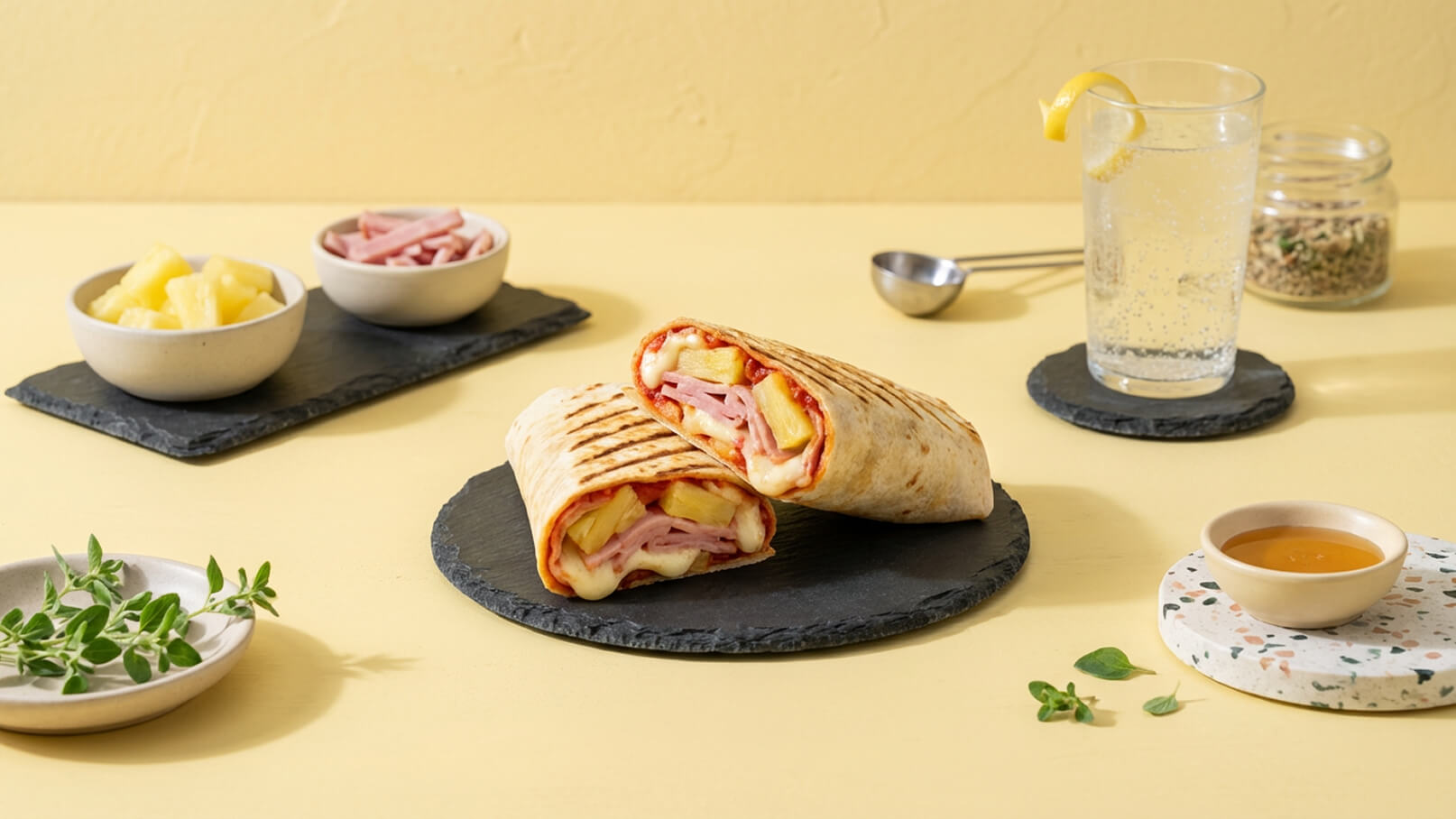 Zwei aufgeschnittene Wraps liegen auf einem schwarzen Teller. Die Füllung besteht aus Käse, Schinken, Ananas und Tomatensoße. Im Hintergrund befinden sich Schüsseln mit Ananasstücken und Schinkenstreifen sowie ein Glas mit Wasser und einer Zitronenscheibe. Der Hintergrund ist gelb.