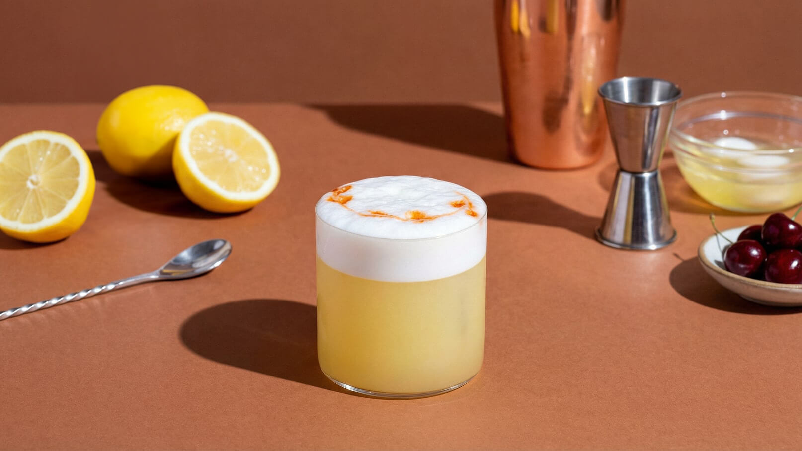 Ein Glas mit einem schaumigen Getränk steht im Vordergrund. Davor liegt ein Löffel, während im Hintergrund Zitronen, ein Messbecher und eine Schüssel mit Kirschen zu sehen sind. Der Tisch hat eine braune Oberfläche.