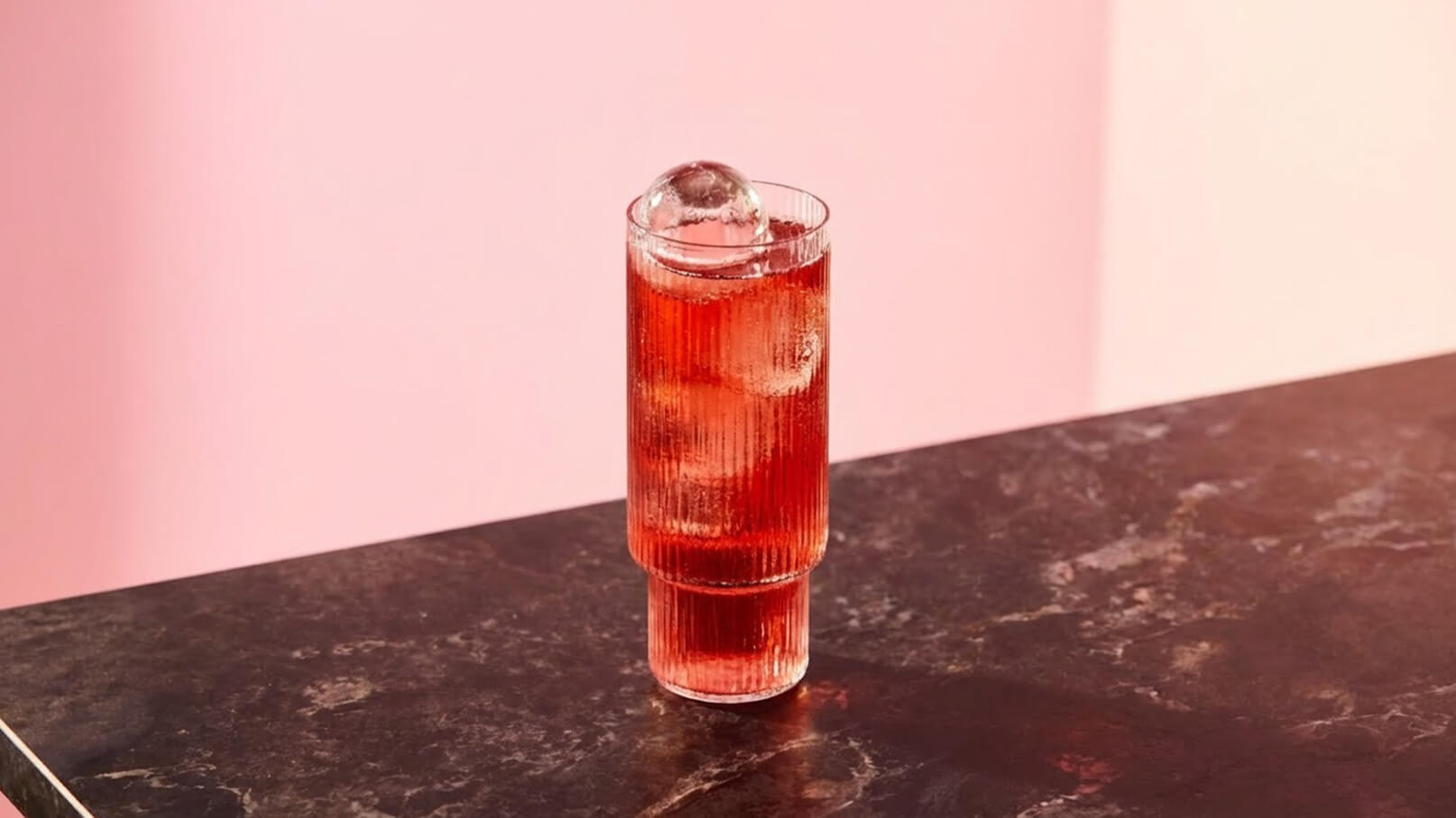 Ein hoher, transparent roter Getränkeglas steht auf einer dunklen marmorierten Oberfläche. Im Glas befindet sich ein runder Eiswürfel. Der Hintergrund ist in sanften, rosa Tönen gehalten.
