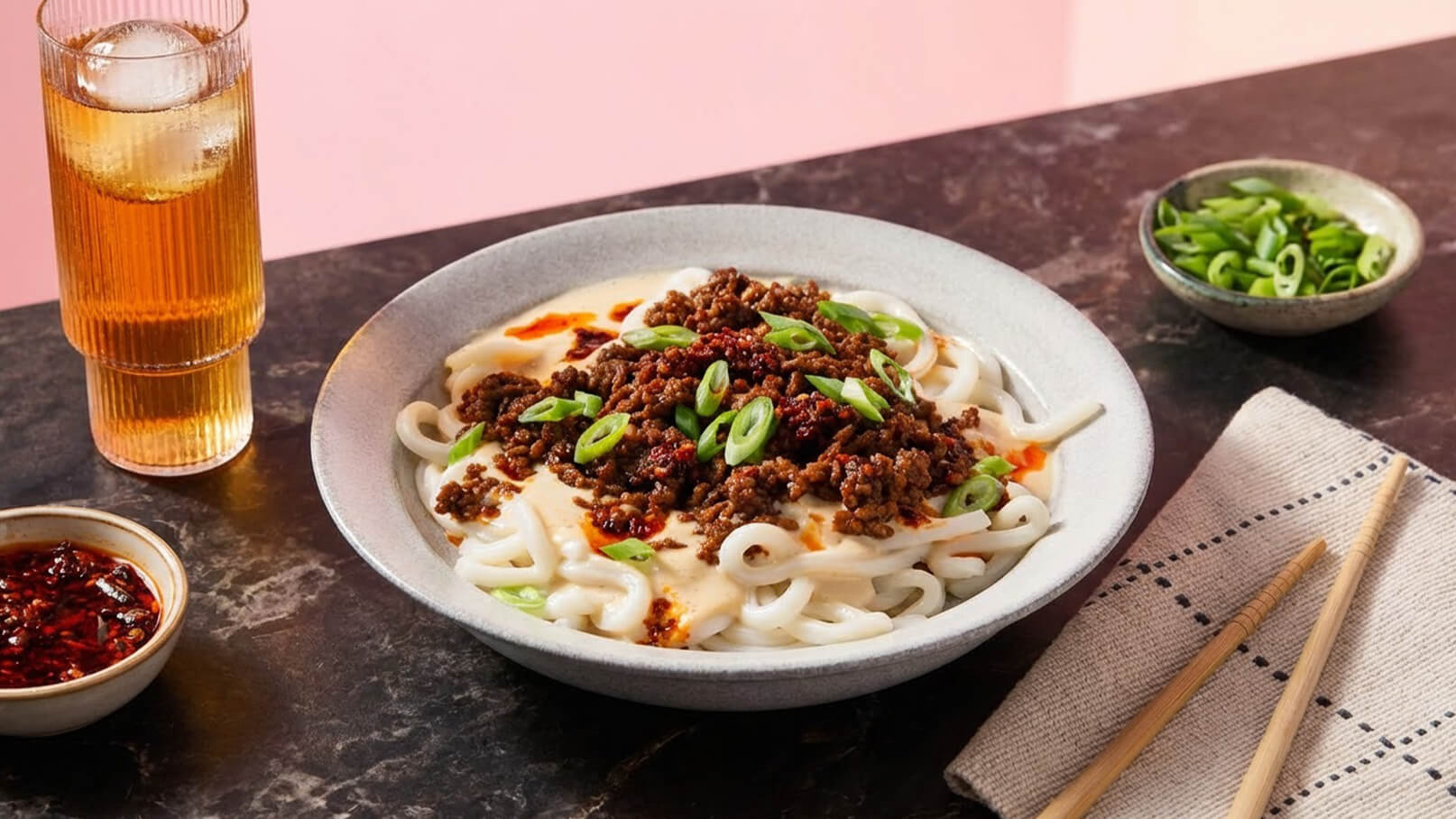 In einer flachen Schale liegen breite Reisnudeln, belegt mit gewürztem Hackfleisch und frischen Frühlingszwiebeln. Neben der Schale stehen kleine Schalen mit Soße und geschnittenen Frühlingszwiebeln sowie ein Glas Tee. Der Hintergrund ist leicht rosa.