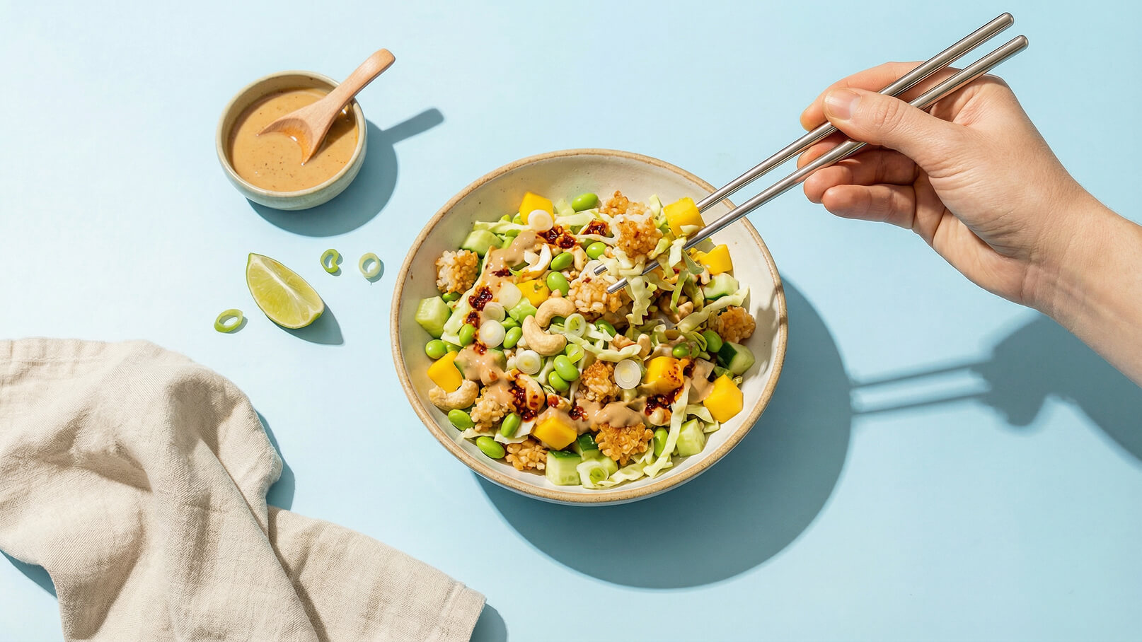 Eine Hand hält Essstäbchen über einer Schüssel mit buntem Salat, der aus Gemüse, Cashews und Mango besteht. Neben der Schüssel befindet sich eine kleine Schale mit Erdnusssauce. Im Hintergrund ist ein blauer Tisch zu sehen.