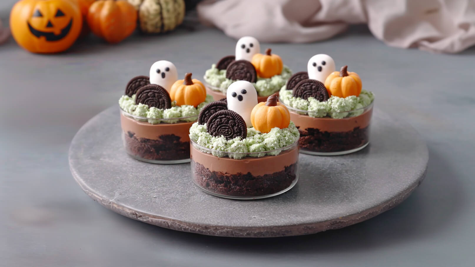 Vier Halloween-Desserts in Gläsern, bestehend aus Schokoladencreme, Keksboden und grüner Sahne. Oben sind kleine Kürbisse aus Marzipan und gruselige Gespenster aus Zucker. Die Desserts stehen auf einer grauen Platte.