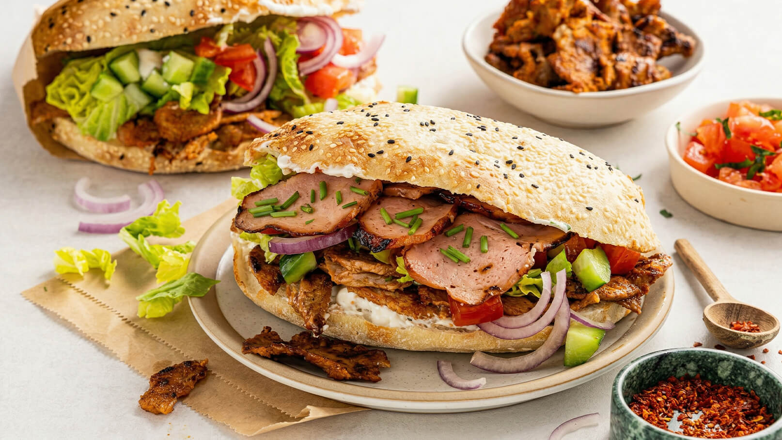 Ein frisches Sandwich mit Sesambrötchen, gefüllt mit Fleisch, geschnittenem Gemüse wie Salat, Gurken und Paprika, sowie Zwiebeln. Im Hintergrund sind weitere Sandwiches und Schalen mit Beilagen und Tomatensalat zu sehen.