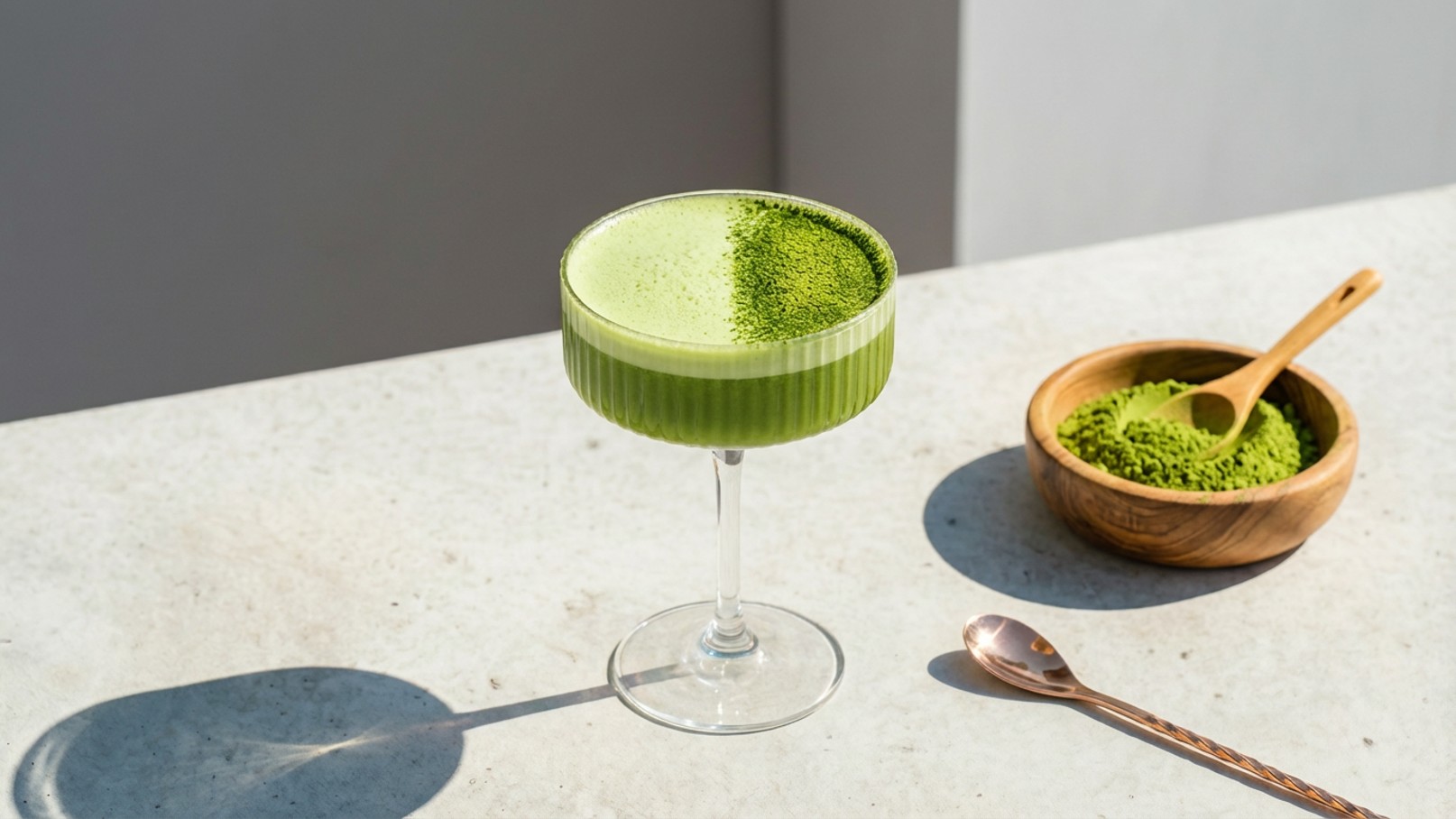 Ein elegantes Glas mit grünem Getränk steht auf einer Tischplatte. Daneben befindet sich eine Schale mit grünem Matcha-Pulver und einem kleinen Löffel. Die Szene vermittelt eine frische, gesunde Atmosphäre.
