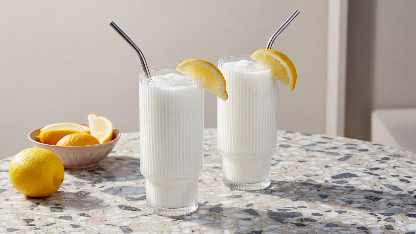 Zwei hohe Gläser mit einem weißen Getränk und einem Strohhalm, jeweils mit einer Zitronenscheibe dekoriert. Im Hintergrund befindet sich eine Schüssel mit frischen Zitronen. Der Tisch hat eine marmorierte Oberfläche.