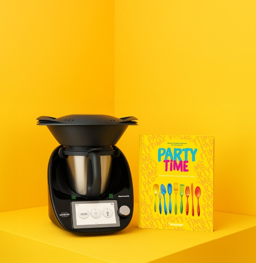 Ein schwarzer Thermomix steht auf einer gelben Oberfläche, daneben liegt ein Kochbuch mit dem Titel „PARTY TIME“. Das Buch zeigt bunte Löffel und Gabeln auf dem Cover und vermittelt das Thema Festtagsgerichte und Rezepte.