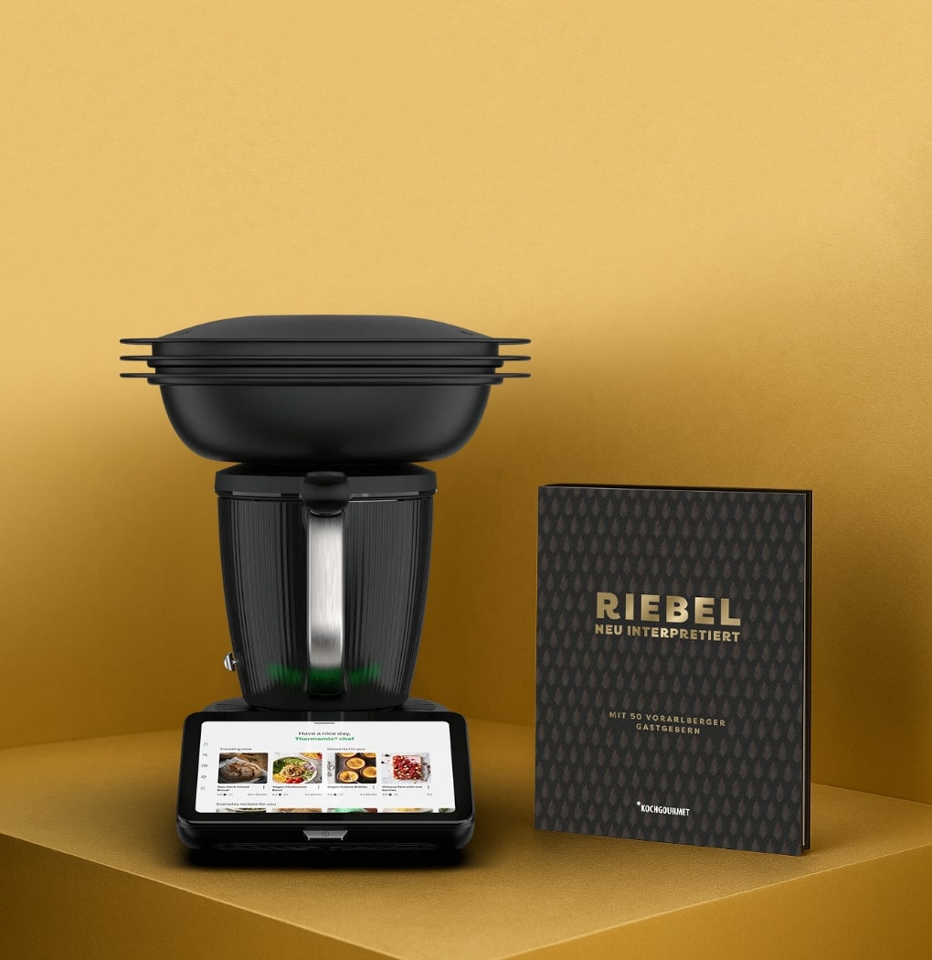 Ein schwarzes Küchengerät mit einem digitalen Display zeigt Rezepte an. Daneben liegt ein Kochbuch mit dem Titel "RIEBEL - NEU INTERPRETIERT". Hintergrund in Gold. Das Bild vermittelt den Fokus auf modernes Kochen und Rezeptideen.