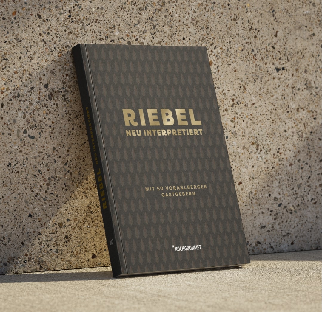 Ein Kochbuch mit dem Titel "RIEBEL - neu interpretiert" liegt auf einer Oberfläche. Der Titel hebt sich in goldenen Buchstaben ab. Es enthält Beiträge von 50 Vorarlberger Gastgebern.