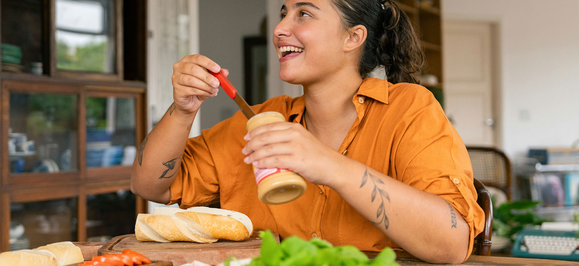 Eine lächelnde Frau in einem orangefarbenen Hemd bereitet ein Sandwich vor. Sie hält ein Messer in der Hand und ist dabei, Erdnussbutter auf ein frisches Baguette zu streichen. Um sie herum liegen frische Zutaten wie Salat und Tomaten.