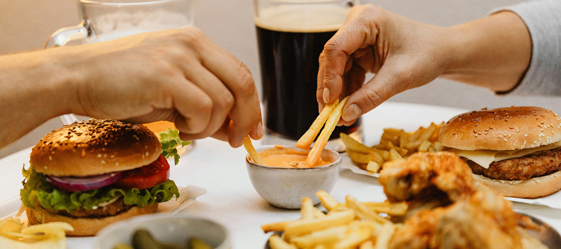 Ein großes Stück Käsepizza wird mit einer Hand gehalten. Auf dem Tisch befindet sich ein Teller mit frittiertem Hähnchen und einer kleinen Schale mit Dip. Daneben steht eine Dose Getränk. Der Hintergrund ist mit einem karierten Tuch dekoriert.